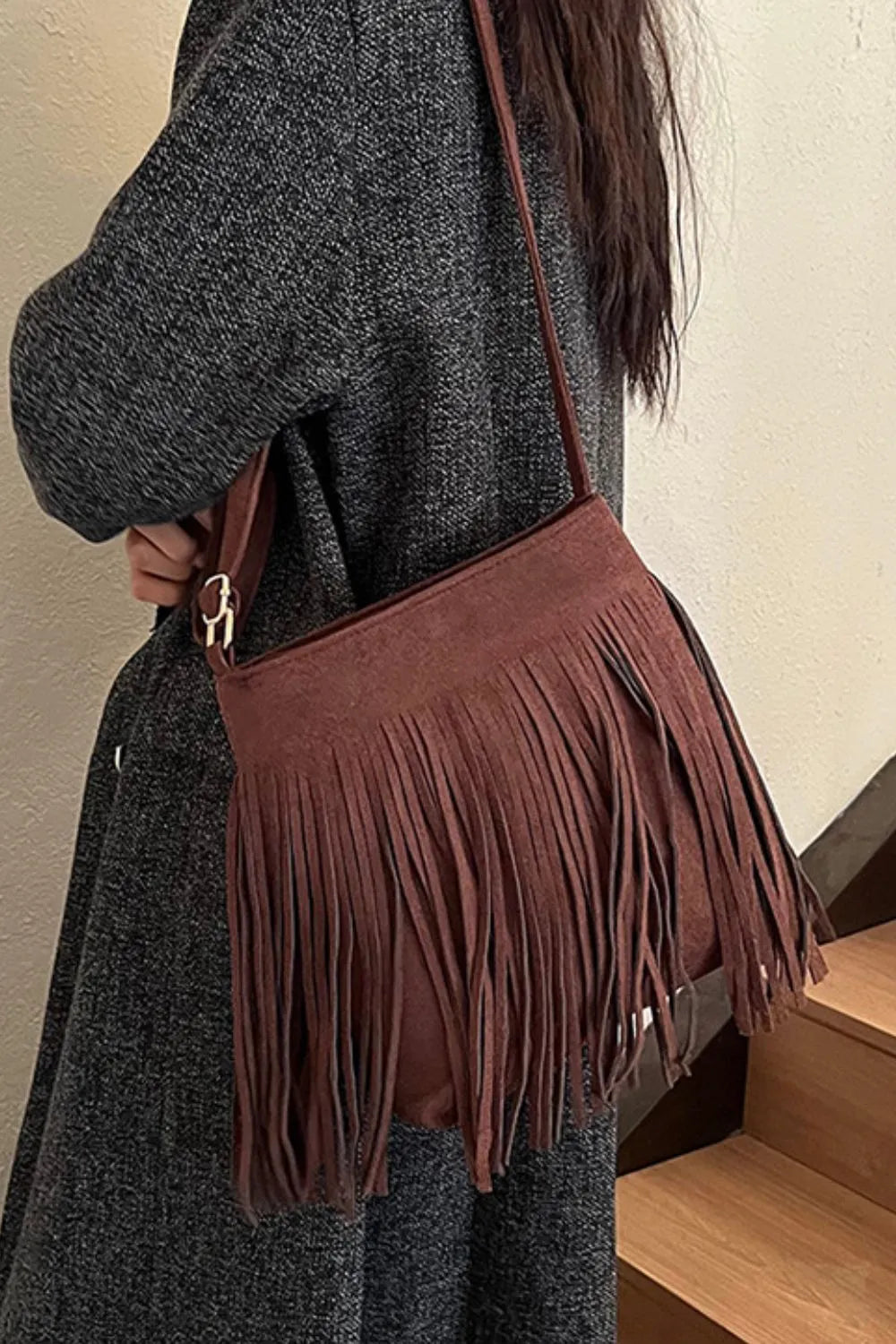 Model wearing a brown fringe shoulder bag with a slim strap over a dark textured coat, side view.