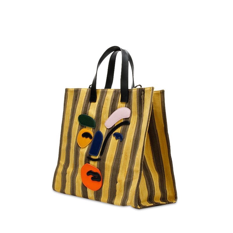 Angled view of a yellow and brown striped tote with black handles and colorful abstract face applique.