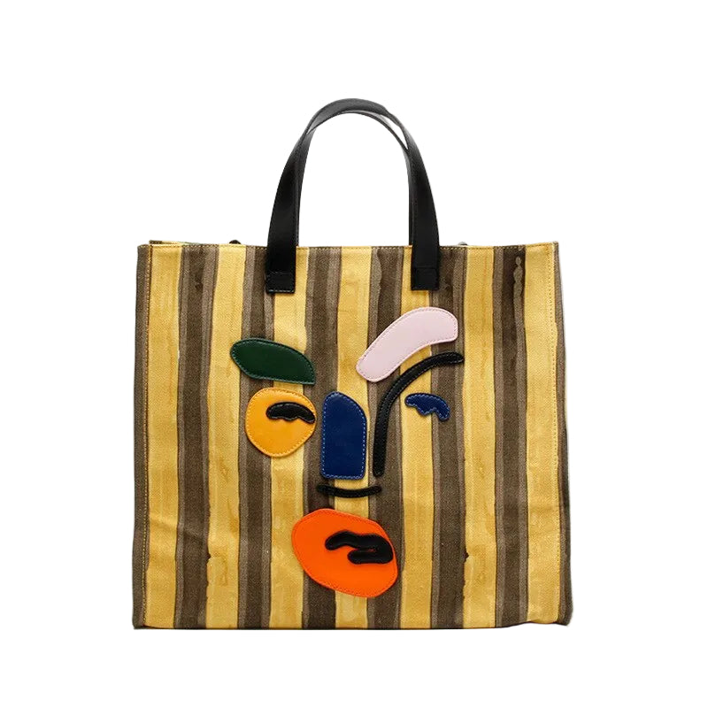 Yellow and brown striped tote with black handles and a colorful abstract face applique, front view.