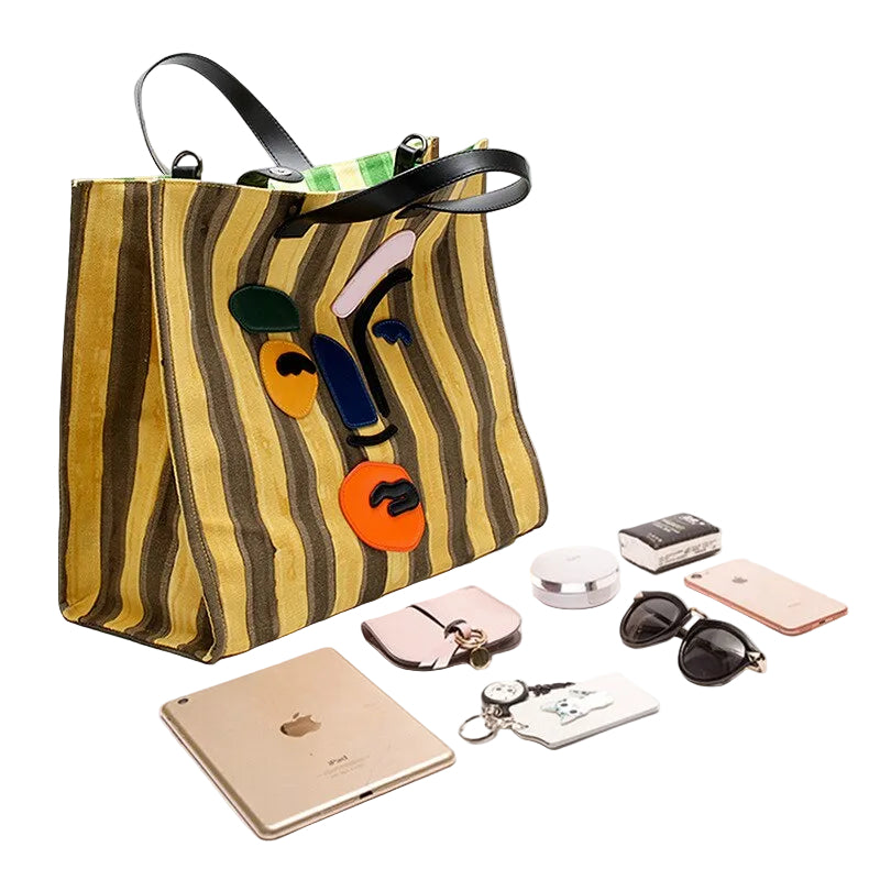 Striped face-applique tote displayed beside a tablet, phone, sunglasses, wallet, keys, and cosmetics.
