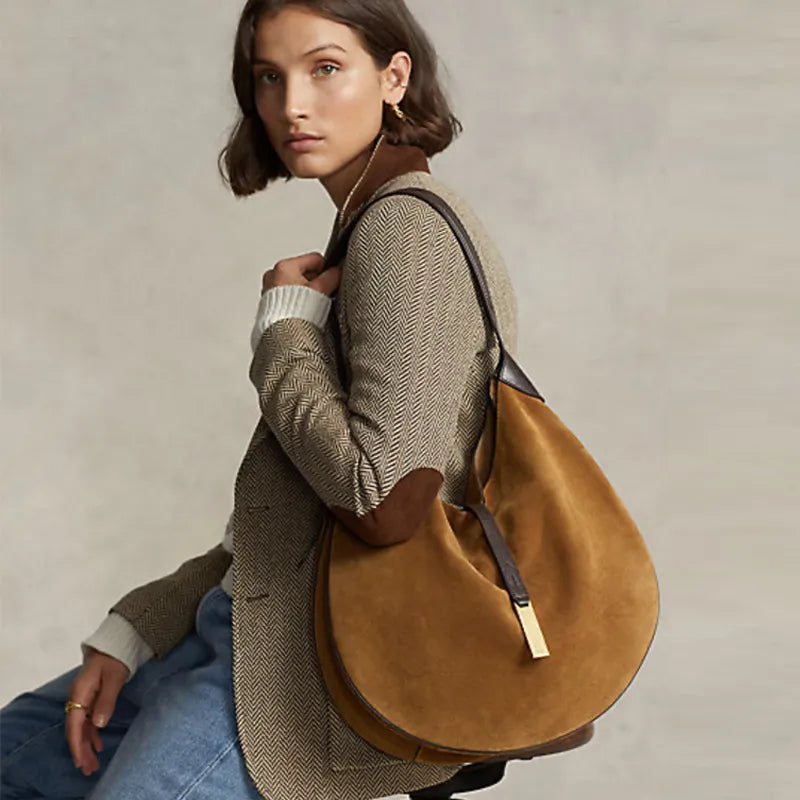 Model wearing a tan rounded shoulder bag with a dark strap and gold-tone front clasp over a checked blazer.