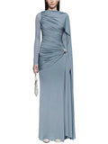Model wearing a dusty blue long-sleeve ruched gown with a draped shoulder panel and silver hip accent, front view.