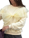 Model wearing a cream sweatshirt with an oversized sheer bow and shoulder ruffles, front view.